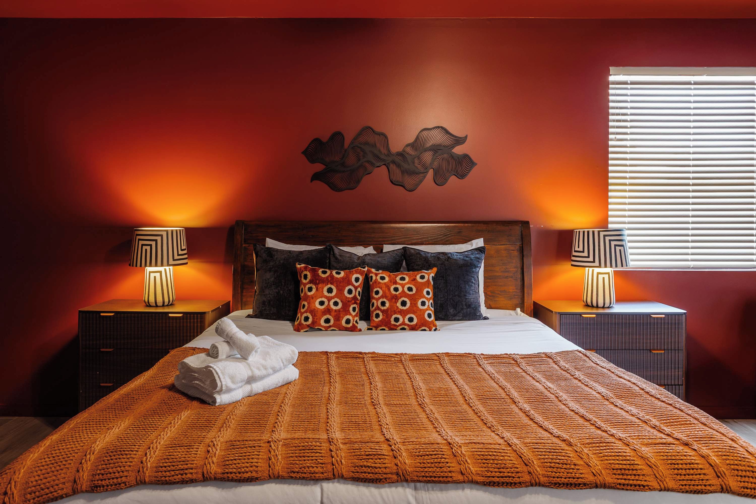 A bedroom with a large bed, two nightstands, a decorative wall piece, and a window with blinds.