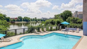 A swimming pool with lounge chairs and umbrellas, surrounded by a fence, with a view of a river and buildings in the background.