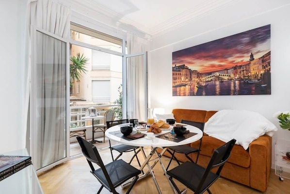 A modern living room with a dining area, a sofa, and a large window with a view of buildings. - Promenade Flat for 3 Guests | 30+ Night Stay (Nice)