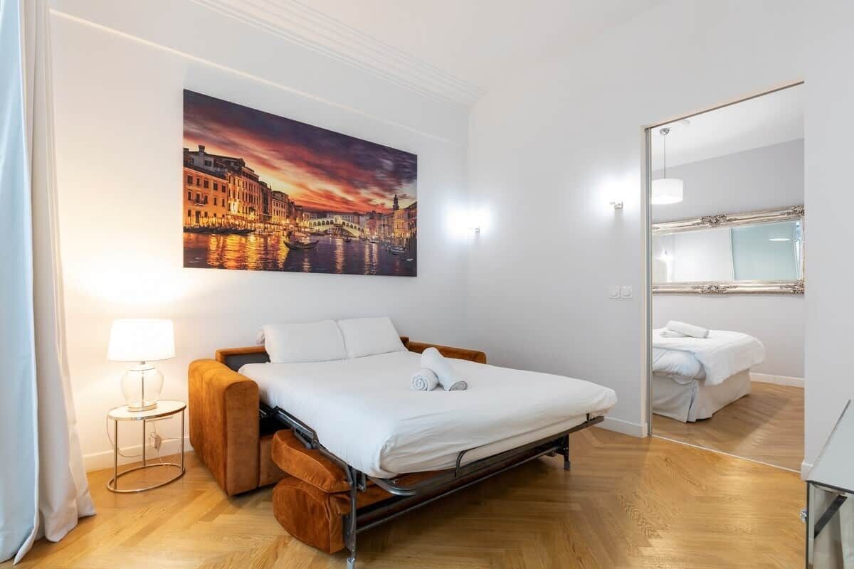 A hotel room with a bed, a bedside table, a lamp, a large painting of a cityscape, and a mirror.