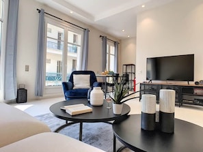 Ein modernes Wohnzimmer mit einer Couch, einem runden Couchtisch, einem blauen Sessel, einem Fernseher und Blick auf einen Balkon.