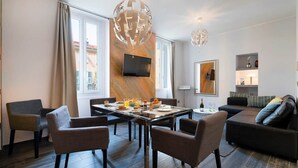 A modern living room with a dining area, a sofa, and a TV mounted on the wall. - Large 3-Room Garibaldi – 4 People | Stay 30+ Days (Nice)