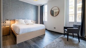 A modern bedroom with a large bed, bedside lamps, a round mirror, a chair, and a view of buildings through the window. - Large 3-Room Garibaldi – 4 People | Stay 30+ Days (Nice)
