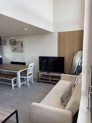 Ein modernes Wohnzimmer mit einer Couch, Fernseher, Esstisch, Stühlen und Pendelleuchten.