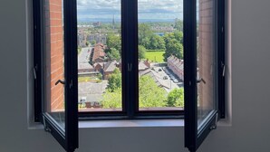 Une pièce dotée d’une grande fenêtre ouverte, d’un mur blanc et offrant une vue sur un paysage urbain avec des bâtiments et de la verdure.