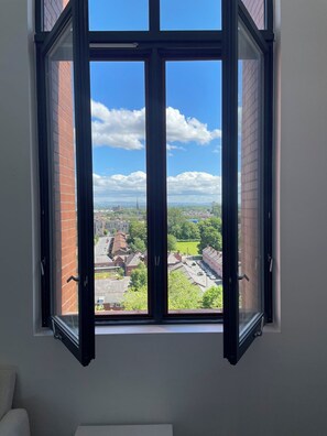 Ein Zimmer mit einem großen offenen Fenster, einer weißen Wand und Blick auf eine Stadtlandschaft mit Gebäuden und Grünflächen.
