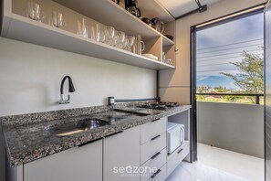 A modern kitchen with granite countertops, a stainless steel sink, and a gas stove. There are shelves above the counter holding glassware and dishes. - Loft 50 m From Camacho Beach Plo0005 (Jaguaruna)