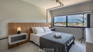 A modern hotel room with a bed, bedside table, lamp, and a view of the city through the window. - Loft 50 m From Camacho Beach Plo0005 (Jaguaruna)