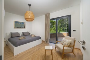 Een slaapkamer met een bed, een stoel, een klein tafeltje, een hanglamp, een schuifdeur met glas en een schilderij aan de muur.