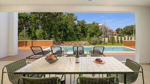 Un espace repas avec une table et des chaises, une piscine en arrière-plan et une vue sur les arbres et les bâtiments.