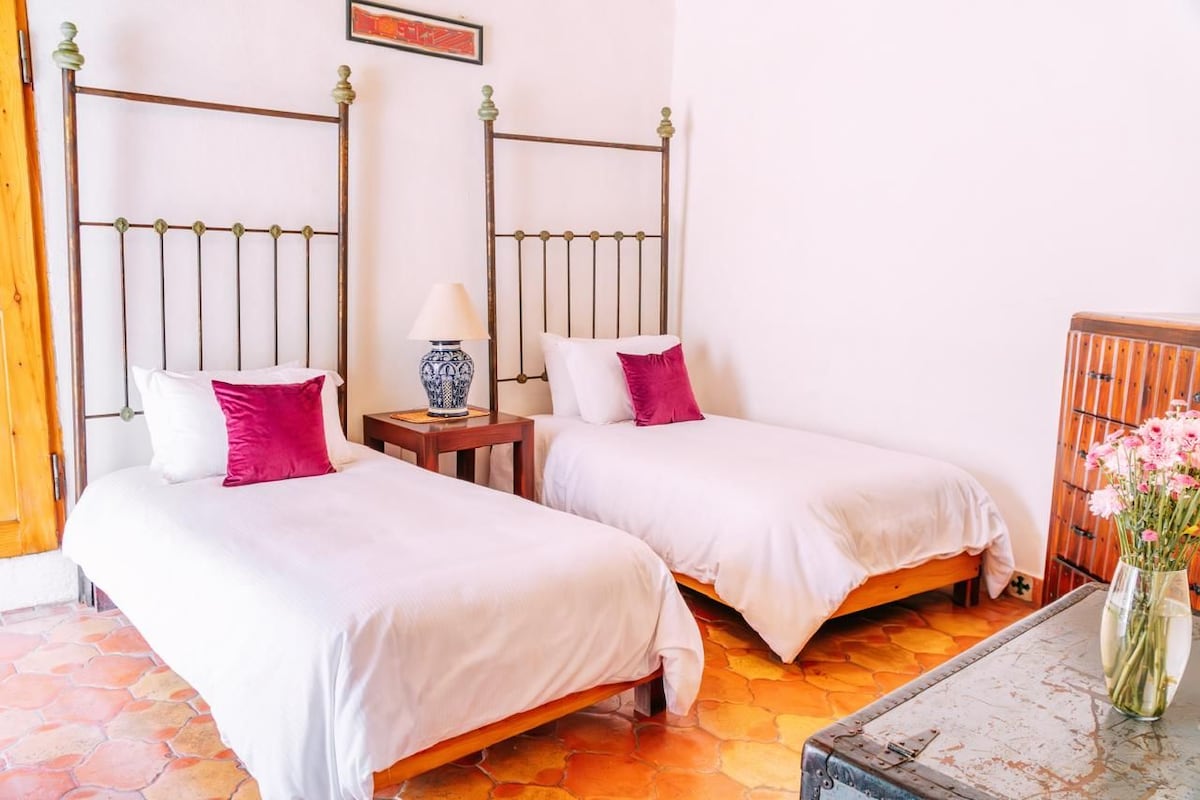 A room with two beds, each with white bedding and a single pink pillow. There is a small table with a lamp and a vase with flowers. The room has a tiled floor and a wooden door.