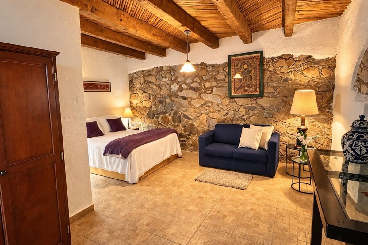 A room with a stone wall, wooden beams, a bed, a sofa, and a table with a vase.
