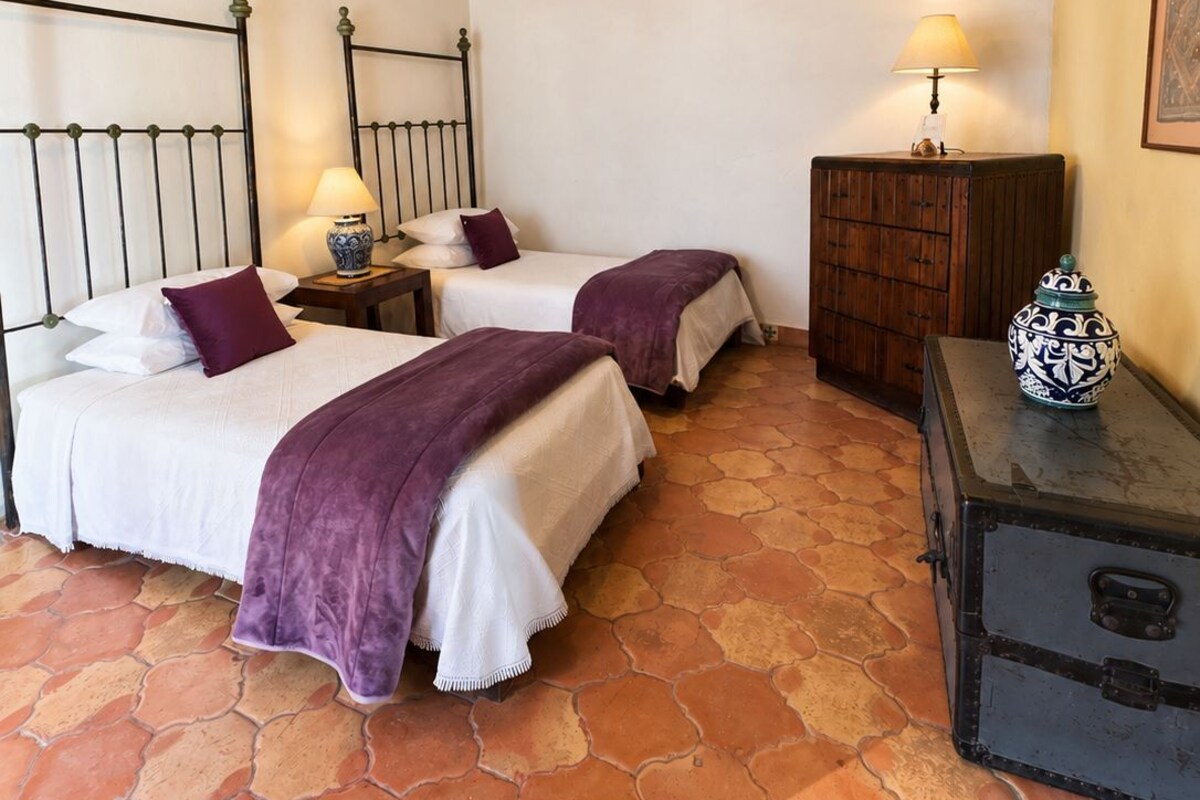 A room with two beds, a wooden ceiling, a tiled floor, a nightstand with a lamp, and a cabinet with a decorative item.