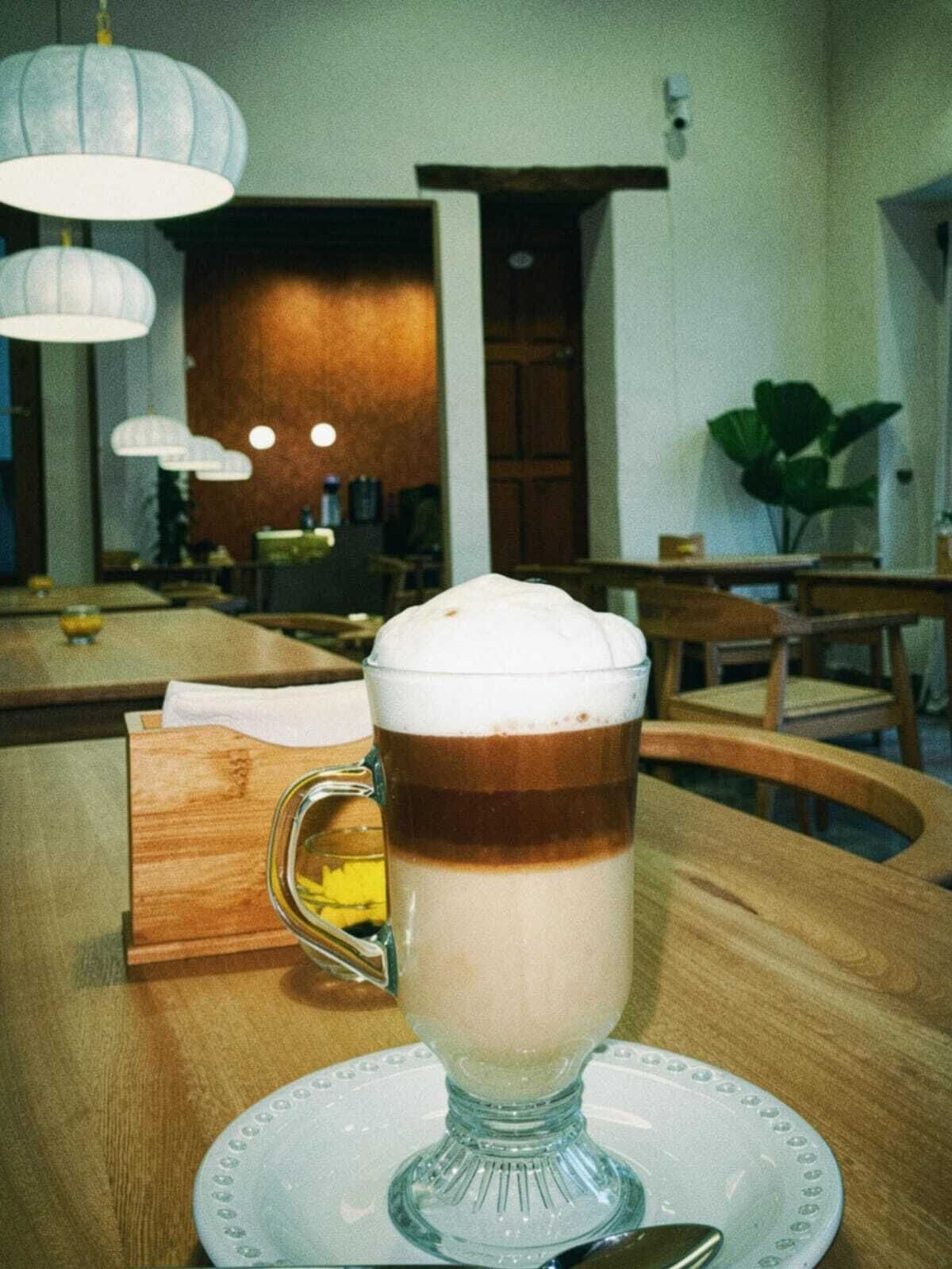 Un vaso de bebida en capas sobre un plato blanco con platillo, sobre una mesa de madera en una habitación con puerta de madera, una planta y luces colgantes.
