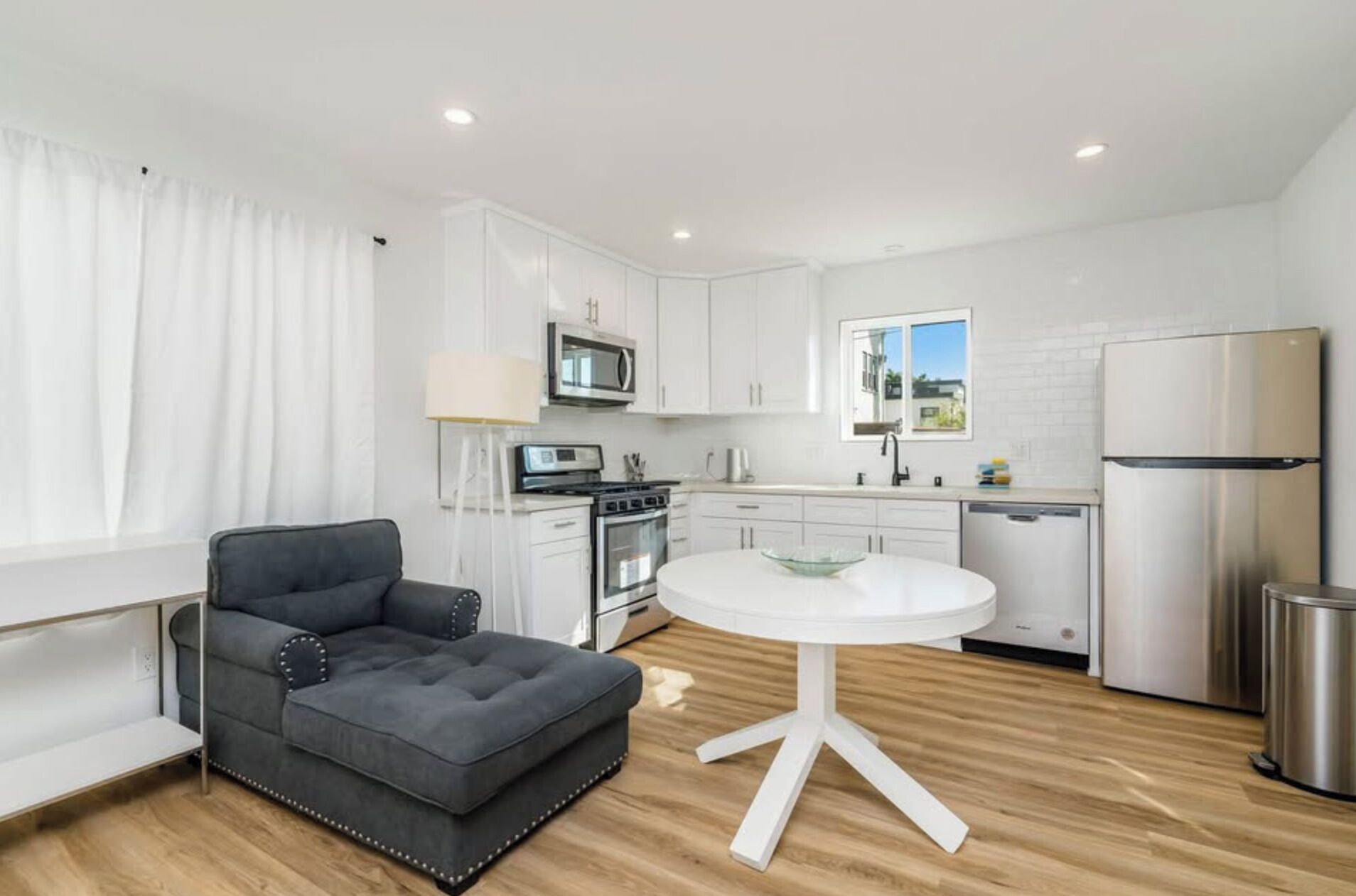 A modern kitchen with white cabinets, stainless steel appliances, a round white table, and a grey sofa.