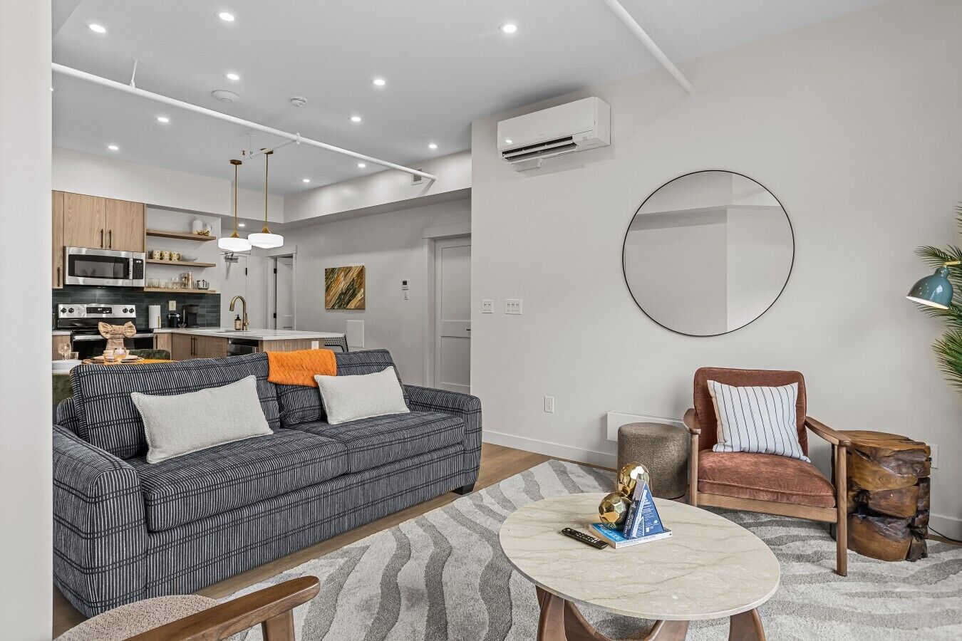 A modern living room with a sofa, armchair, and coffee table, a kitchen area with appliances, and a large mirror on the wall.