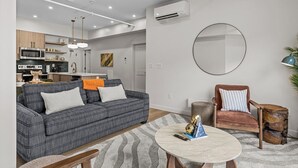 Ein modernes Wohnzimmer mit Sofa, Sessel und Couchtisch, ein Küchenbereich mit Geräten sowie ein großer Spiegel an der Wand.