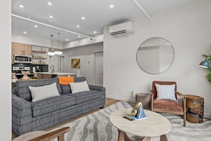 En moderne stue med sofa, lænestol og sofabord, et køkken med hårde hvidevarer, og et stort spejl på væggen.