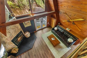 Ein Interieur einer Hütte mit Kamin aus Stein, einer Couch, Holzboden und Blick auf den Wald durch ein großes Fenster.