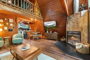Ein gemütliches Blockhaus-Interieur mit Kamin aus Naturstein, Holzwänden, einem Essbereich und einer durch einen offenen Durchgang sichtbaren Küche.