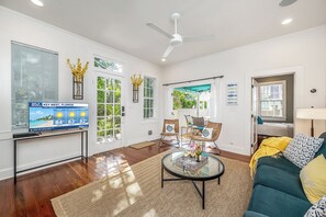 A spacious living room with a TV displaying a weather forecast, a ceiling fan, and a view of the outdoors through large windows. - Reynolds Paradise | Walk to Higgs Beach (Key West)