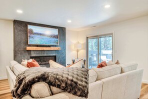A living room with a fireplace, a television mounted on a stone wall, and a large window. - Pine & Powder - SHARC, Hot Tub, Bikes, EV Charger (Sunriver)
