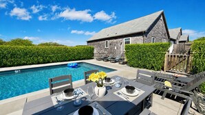 Un área de comedor junto a la piscina con una mesa lista para dos personas, una piscina y una casa de madera al fondo.