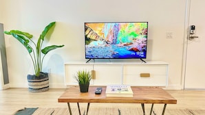 A living room with a television on a white cabinet, a wooden coffee table with a remote and a magazine, a potted plant, and a striped rug. - Broadway (San Diego)