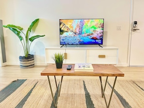 A living room with a television on a white cabinet, a wooden coffee table with a remote and a magazine, a potted plant, and a striped rug. - Broadway (San Diego)