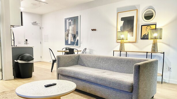 A modern living room with a grey sofa, a round coffee table, and a dining area with a small table and chairs. - Broadway (San Diego)