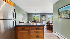A kitchen with a large island, wooden cabinets, and a refrigerator. There is a dining area with a table and chairs, and a sofa. - Lies by Avantstay Indoor Heated Pool + Game Lounge Pool Table + Foosball (North Creek)