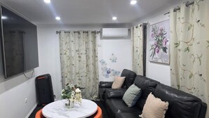 A living room with a black leather sofa, a round marble table with a vase of flowers, a flat-screen TV mounted on the wall, and floral curtains.