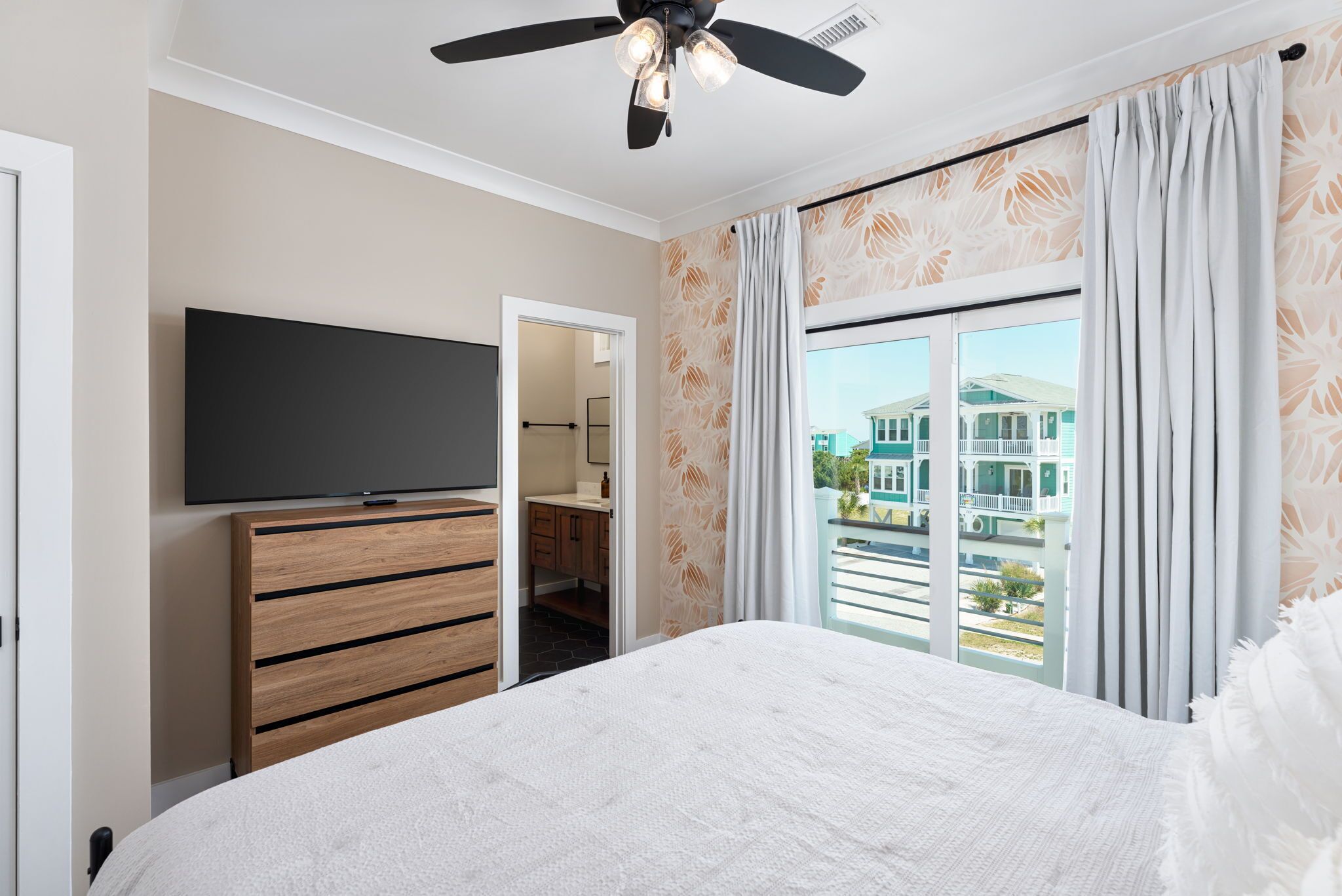 A bedroom with a bed, a TV on a wooden dresser, a ceiling fan, and a view of a building through a window.