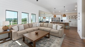 Ein modernes Wohnzimmer mit einem Ecksofa, einem Couchtisch und einem Essbereich, im Hintergrund ist eine Küche zu sehen.