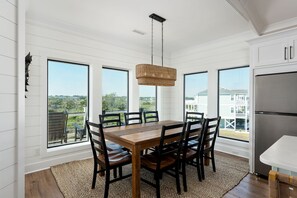 Un comedor con una mesa de madera, sillas negras, una lámpara colgante tejida y ventanas amplias que ofrecen vistas a un edificio blanco y follaje.