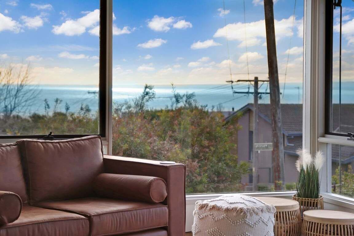 Una habitación con un sofá de cuero marrón, un pouf redondo y vistas al mar a través de amplios ventanales.