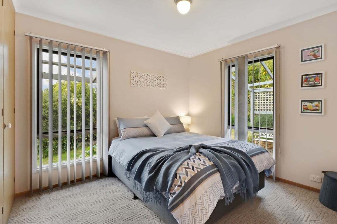 A bedroom with a bed, a window with blinds, a wall-mounted light, and a view of greenery outside.