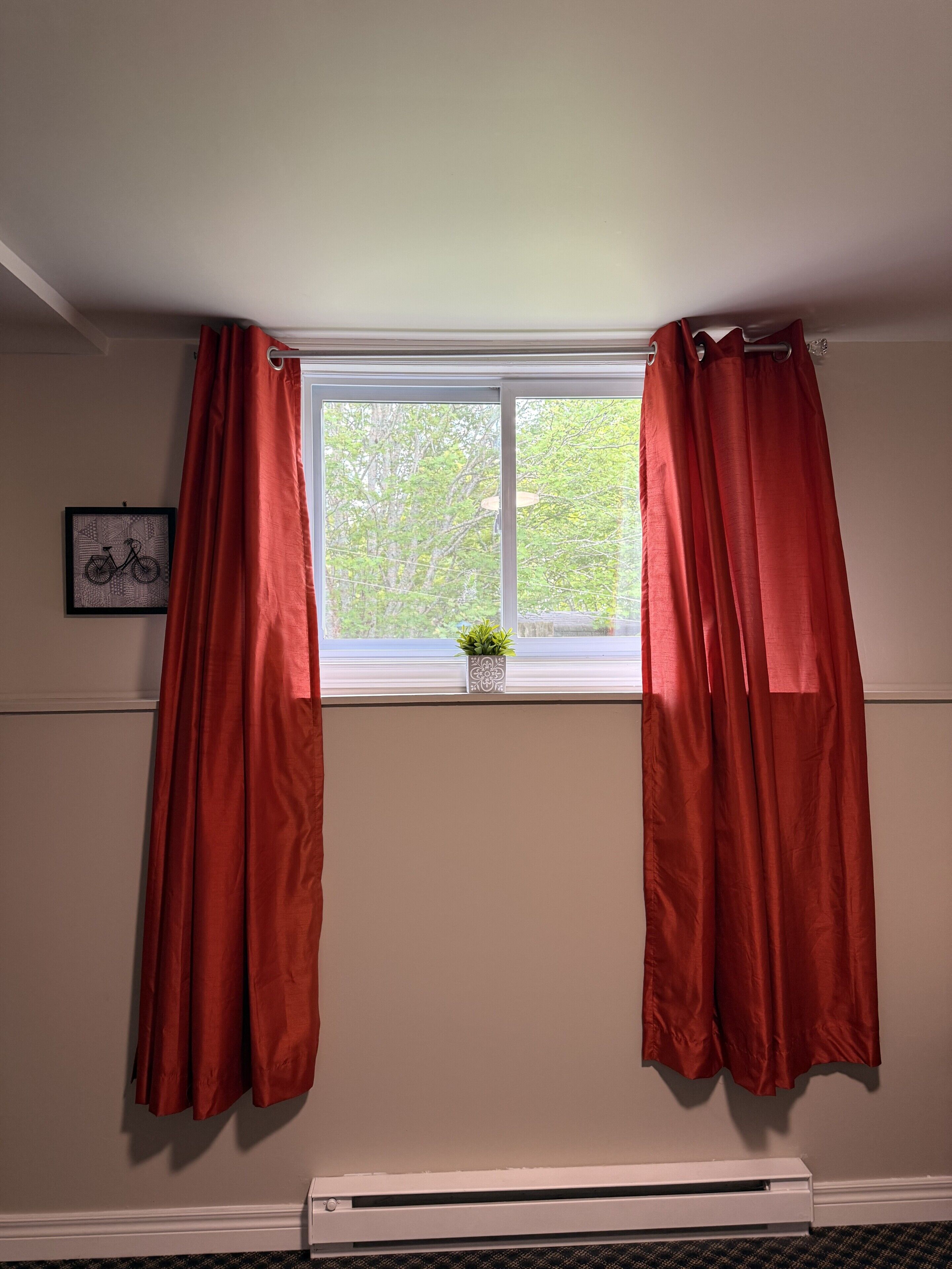Une pièce avec une fenêtre, des rideaux rouges, une petite plante sur le rebord de la fenêtre et un tableau encadré au mur.