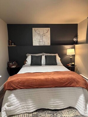 Une chambre à coucher comprenant un lit, une tête de lit, une œuvre d’art accrochée au mur, une lampe de chevet et une petite table avec une lampe.