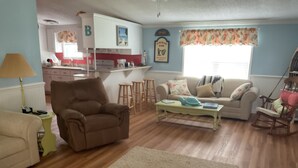 A living room with a brown armchair, a white sofa, a coffee table, and a rocking chair. There is a kitchen in the background with a B-shaped letter decoration. - Shellpoint in Townsend Ga. Spacious 3-bedroom mobile home lodge fee 129. a night (Townsend)