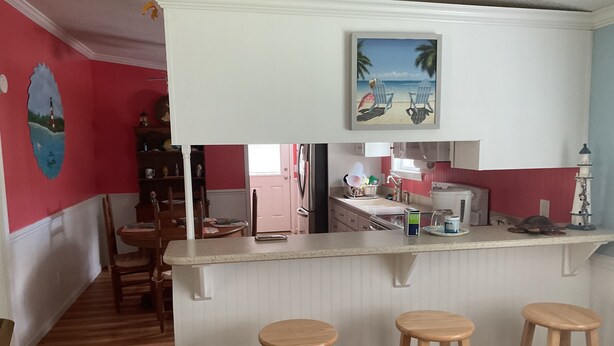 A kitchen with a bar area, wooden stools, and a painting of a beach scene. - Shellpoint in Townsend Ga. Spacious 3-bedroom mobile home lodge fee 129. a night (Townsend)