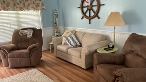 A living room with a beige sofa, two brown armchairs, a wooden floor, a white rug, a ship's wheel decoration, a lamp, and a small table with a remote control. - Shellpoint in Townsend Ga. Spacious 3-bedroom mobile home lodge fee 129. a night (Townsend)