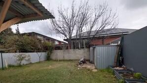 Een achtertuin met een gazon, een betonnen muur, een metalen tuinhuisje en een boom.