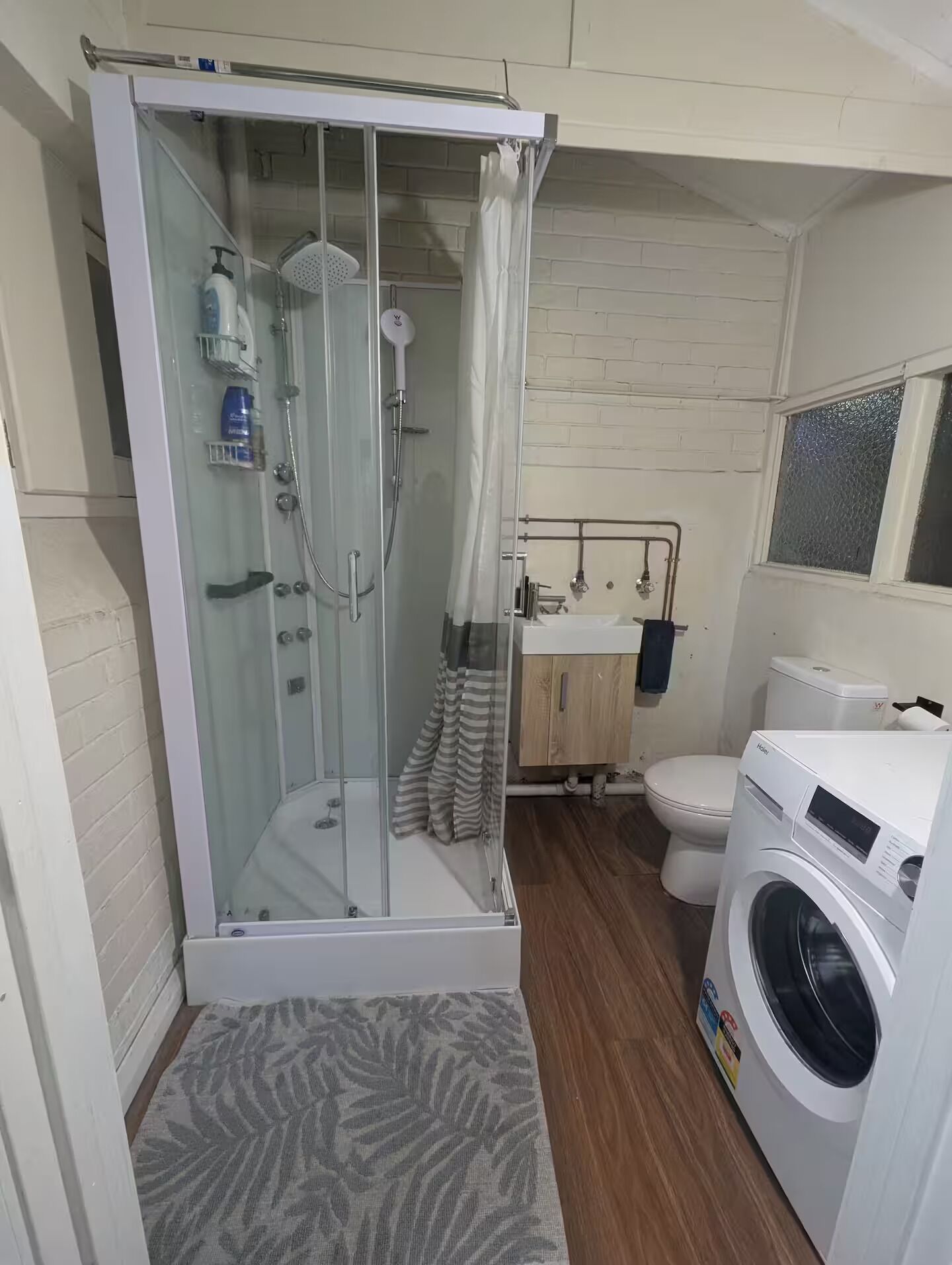 Et badeværelse med en glasafskærmet brusekabine, toilet, vaskemaskine og et spejl over vasken.