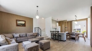 Ein modernes Wohnzimmer mit einem Ecksofa, Hocker, einem Essbereich mit Stühlen, einer Küche mit Bar und einer Treppe.