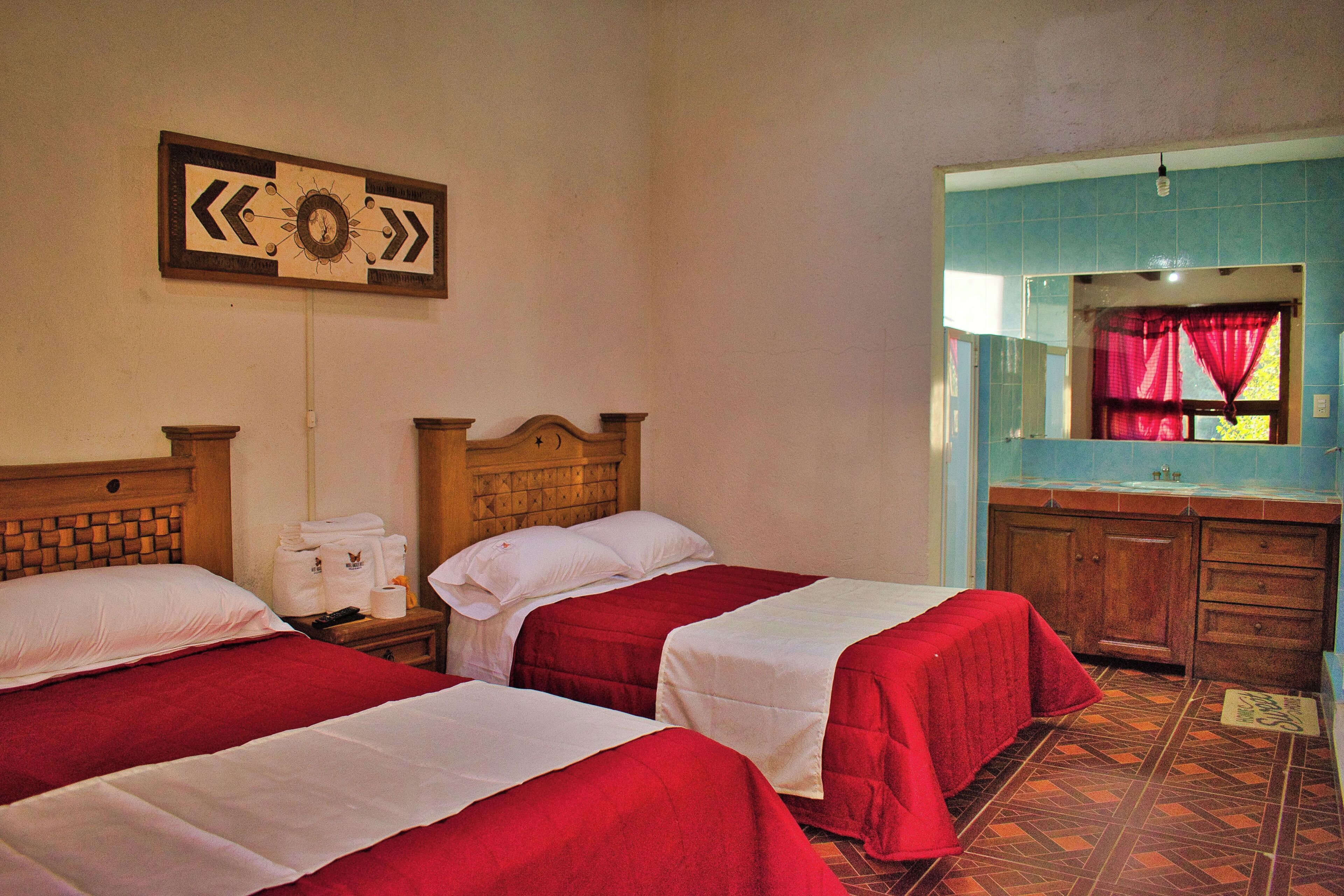 Une chambre d’hôtel avec deux lits, une tête de lit en bois, un miroir, un lavabo et une fenêtre avec un rideau rouge.