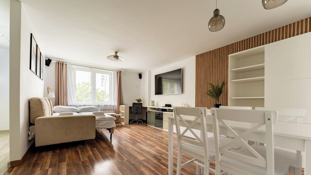 A modern living room with a sofa, a dining table with chairs, a TV mounted on the wall, and a wooden floor. - Gdynia Marina Harmony Apartment (Gdynia)