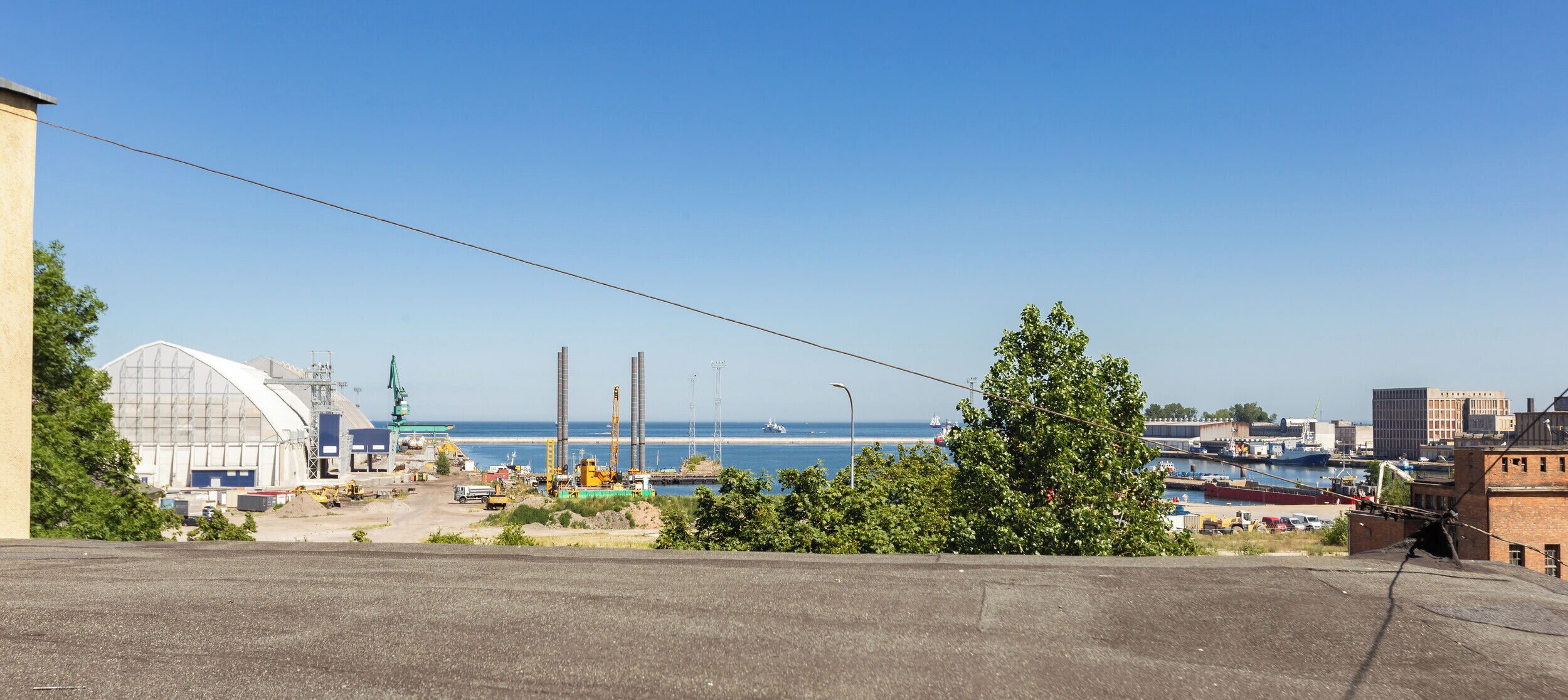 Ein Küstenindustriepark mit Baufahrzeugen, einem großen Gebäude mit gewölbtem Dach und freiem Blick auf das Meer.