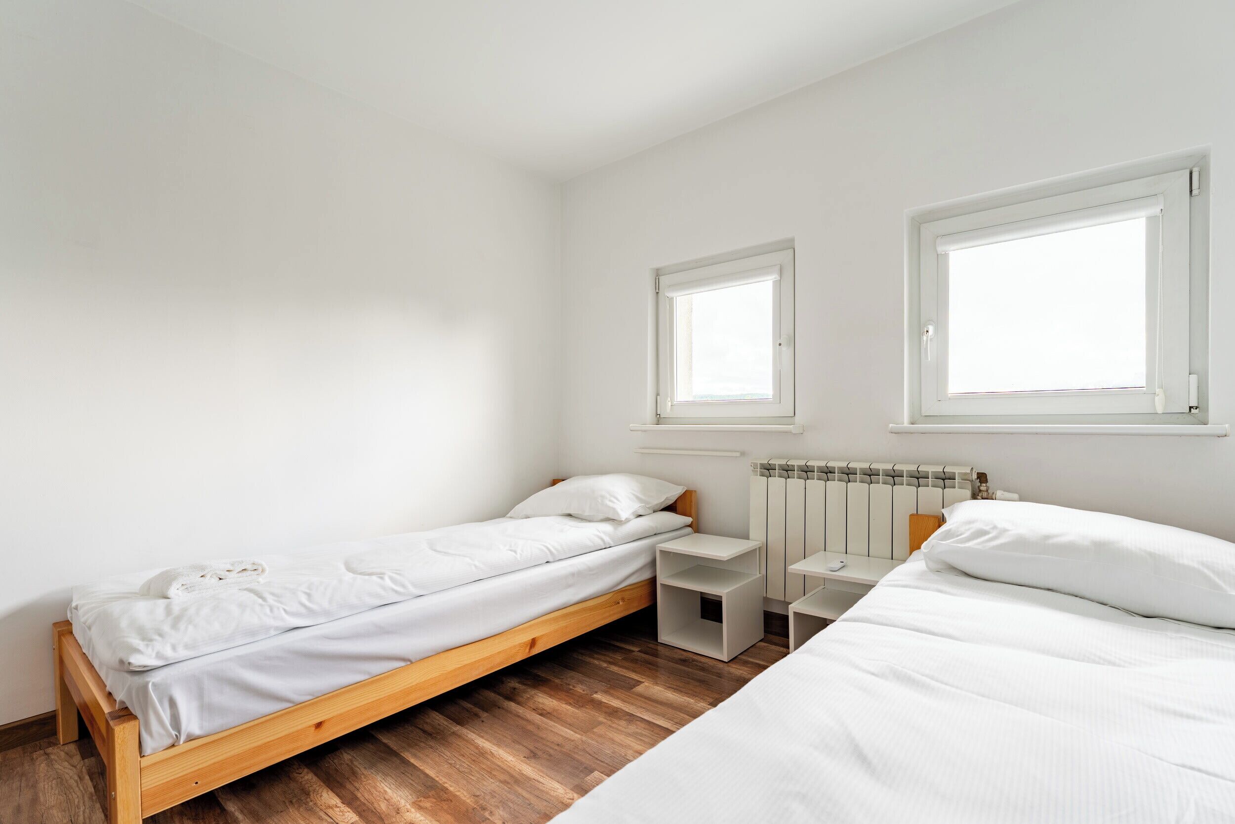 Una habitación pequeña con cama individual, piso de madera, paredes blancas y dos ventanas.