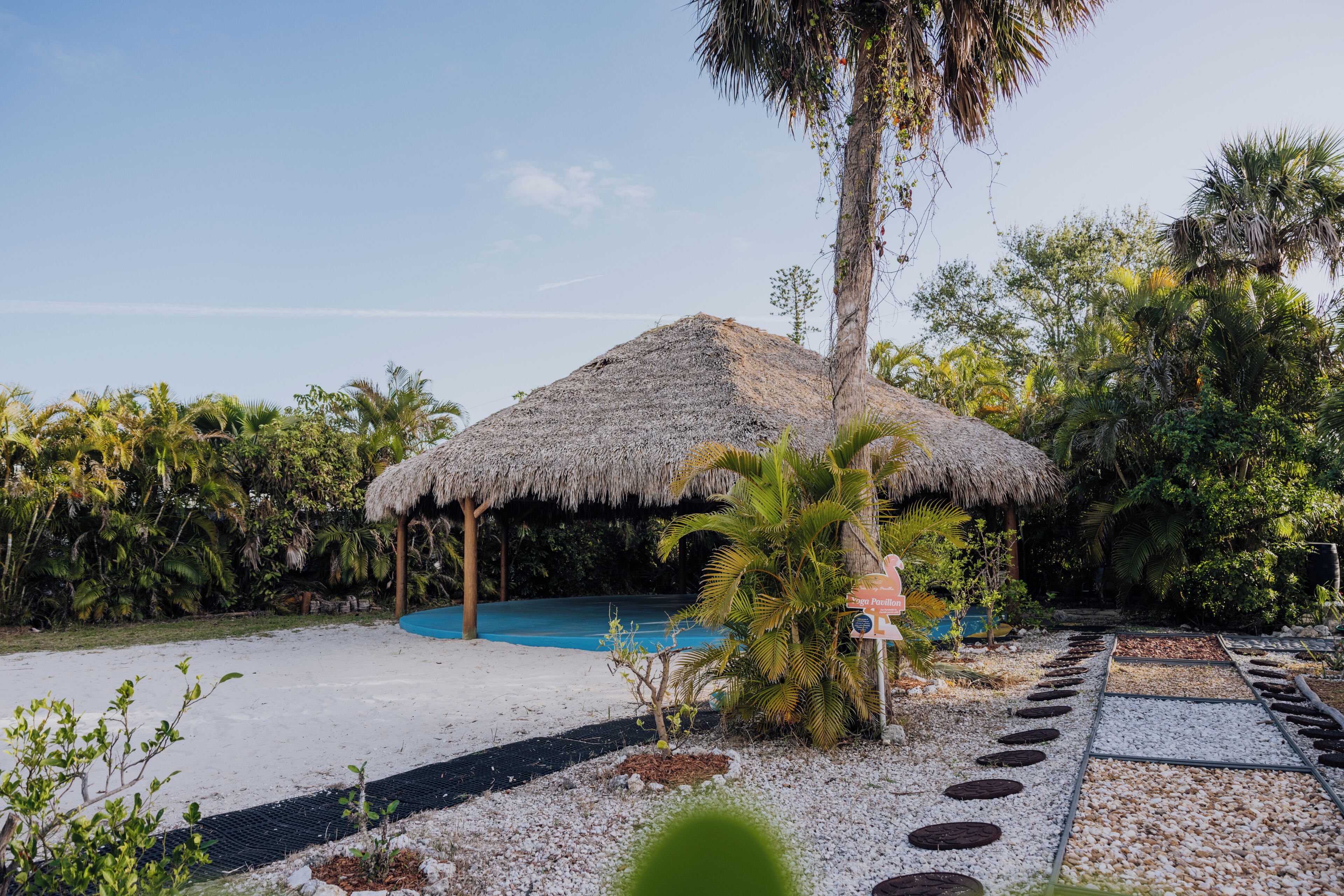 A thatched-roof structure with a pool, surrounded by tropical vegetation and a pebble path.